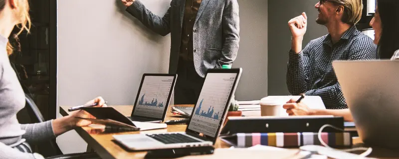 Personnes en réunion dans un bureau, discutant communication devant un tableau blanc. Plusieurs ordinateurs portables montrent des graphiques, suggérant une présentation ou une analyse de données.